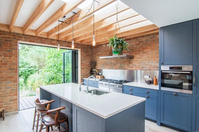 Inspiration for a medium sized modern galley open plan kitchen in Surrey with shaker cabinets, blue cabinets, metallic splashback, stainless steel appliances, an island, an integrated sink, engineered stone countertops, brick splashback, light hardwood flooring and white worktops.
