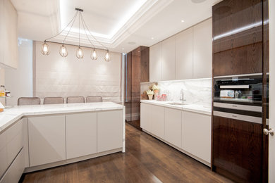 This is an example of a contemporary l-shaped eat-in kitchen in London with a drop-in sink, flat-panel cabinets, white cabinets, marble benchtops, white splashback, marble splashback, stainless steel appliances, a peninsula and brown floor.