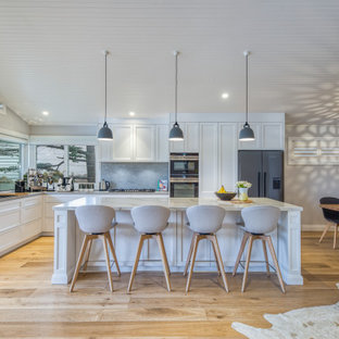 Design ideas for a large beach style l-shaped kitchen in Sydney with a drop-in sink, shaker cabinets, white cabinets, grey splashback, stainless steel appliances, light hardwood floors, with island, beige floor and white benchtop.