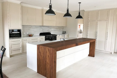 Photo of a mid-sized traditional galley open plan kitchen in Melbourne with a farmhouse sink, shaker cabinets, beige cabinets, white splashback, porcelain splashback, black appliances, porcelain floors, with island and wood benchtops.