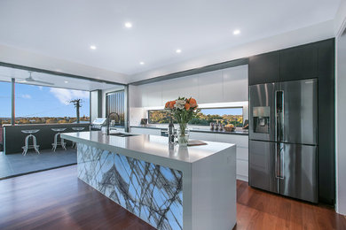 Large contemporary single-wall open plan kitchen in Brisbane with a double-bowl sink, flat-panel cabinets, white cabinets, black appliances, medium hardwood floors, brown floor, white benchtop, quartz benchtops, white splashback, glass sheet splashback and with island.