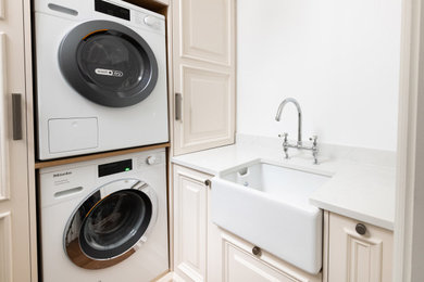 Inspiration for a small victorian l-shaped utility room in London with a belfast sink, shaker cabinets, beige cabinets, engineered stone countertops, white walls, dark hardwood flooring, a stacked washer and dryer, brown floors and white worktops.