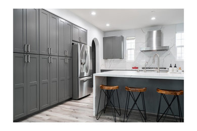 Mid-sized minimalist l-shaped light wood floor, multicolored floor and vaulted ceiling eat-in kitchen photo in Chicago with a drop-in sink, raised-panel cabinets, gray cabinets, granite countertops, white backsplash, granite backsplash, stainless steel appliances, an island and white countertops