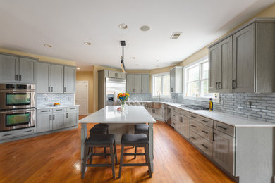 Inspiration for a large transitional u-shaped medium tone wood floor and brown floor eat-in kitchen remodel in Other with a farmhouse sink, shaker cabinets, gray cabinets, quartz countertops, gray backsplash, glass tile backsplash, stainless steel appliances, an island and white countertops
