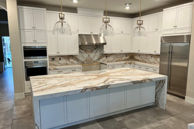 Example of a large transitional l-shaped porcelain tile and gray floor eat-in kitchen design in Orange County with a farmhouse sink, shaker cabinets, white cabinets, white backsplash, stainless steel appliances, an island and white countertops