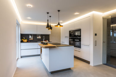 Design ideas for a mid-sized contemporary l-shaped kitchen in Frankfurt with white cabinets, black appliances, with island, a single-bowl sink, flat-panel cabinets, black splashback, concrete floors, grey floor and black benchtop.
