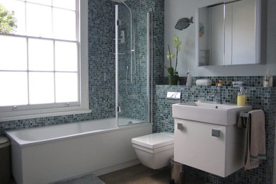 Mid-sized modern master bathroom in London with a wall-mount sink, flat-panel cabinets, white cabinets, a drop-in tub, a shower/bathtub combo, a wall-mount toilet, blue tile, mosaic tile, white walls and light hardwood floors.