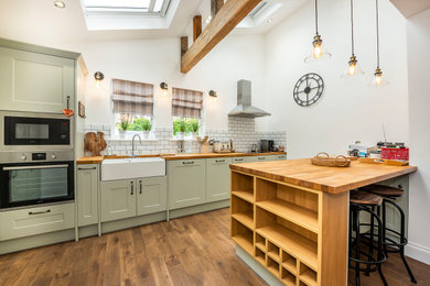 Inspiration for a medium sized classic open plan kitchen in London with a single-bowl sink, shaker cabinets, green cabinets, wood worktops, white splashback, metro tiled splashback, stainless steel appliances, medium hardwood flooring, an island, brown floors, brown worktops, exposed beams and a chimney breast.