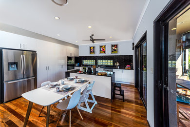 This is an example of a modern l-shaped eat-in kitchen in Gold Coast - Tweed with a double-bowl sink, flat-panel cabinets, white cabinets, quartz benchtops, black splashback, subway tile splashback, stainless steel appliances, medium hardwood floors, with island and brown floor.