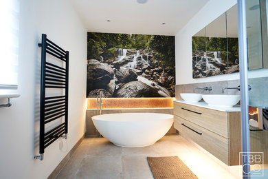 Medium sized contemporary ensuite bathroom in Sussex with flat-panel cabinets, light wood cabinets, a freestanding bath, a corner shower, a wall mounted toilet, grey tiles, porcelain tiles, white walls, porcelain flooring, a vessel sink, quartz worktops, grey floors, a hinged door and white worktops.