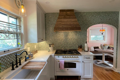 Example of a small french country light wood floor enclosed kitchen design in Kansas City with a farmhouse sink, shaker cabinets, white cabinets, quartz countertops, green backsplash, glass sheet backsplash, stainless steel appliances and white countertops