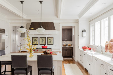 Traditional u-shaped eat-in kitchen in Boston with a farmhouse sink, recessed-panel cabinets, white cabinets, white splashback, stainless steel appliances, medium hardwood floors, with island, brown floor and white benchtop.