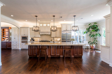 Photo of a large transitional single-wall eat-in kitchen in Dallas with a farmhouse sink, raised-panel cabinets, white cabinets, granite benchtops, beige splashback, ceramic splashback, stainless steel appliances, dark hardwood floors, with island and brown floor.