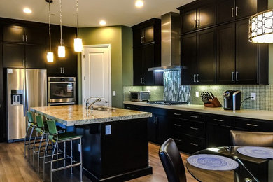Large minimalist single-wall ceramic tile open concept kitchen photo in Las Vegas with an undermount sink, brown cabinets, granite countertops, green backsplash, glass sheet backsplash, stainless steel appliances and an island