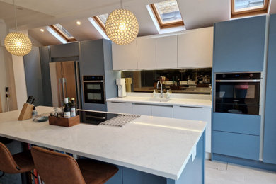 Photo of a large contemporary galley open plan kitchen in London with a built-in sink, flat-panel cabinets, blue cabinets, quartz worktops, metallic splashback, glass sheet splashback, black appliances, porcelain flooring, an island, white floors, white worktops and a vaulted ceiling.
