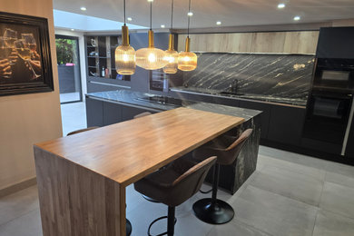 This is an example of a large contemporary grey and black l-shaped kitchen/diner in West Midlands with an integrated sink, flat-panel cabinets, black cabinets, quartz worktops, black splashback, granite splashback, black appliances, porcelain flooring, an island, grey floors, black worktops, a coffered ceiling and a feature wall.