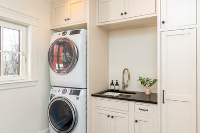 Laundry room - transitional porcelain tile and black floor laundry room idea in Boston with an undermount sink, shaker cabinets, beige cabinets, beige walls, a stacked washer/dryer and black countertops
