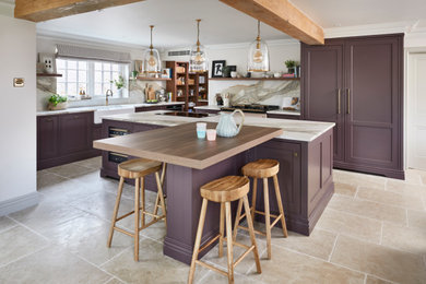 Medium sized classic open plan kitchen in Berkshire with a belfast sink, beaded cabinets, purple cabinets, marble worktops, white splashback, marble splashback, black appliances, porcelain flooring, an island, beige floors, white worktops, exposed beams and a feature wall.
