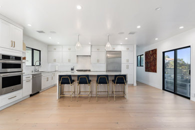 Minimalist u-shaped medium tone wood floor and brown floor kitchen photo in Los Angeles with a double-bowl sink, shaker cabinets, white cabinets, quartzite countertops, white backsplash, stainless steel appliances, an island and multicolored countertops