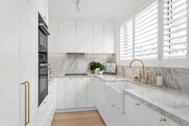 Mid-sized traditional galley separate kitchen in Sydney with a farmhouse sink, shaker cabinets, white cabinets, glass benchtops, grey splashback, porcelain splashback, black appliances, light hardwood floors, no island, beige floor and grey benchtop.