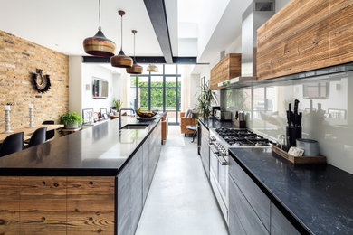 Mid-sized contemporary galley open plan kitchen in London with a drop-in sink, flat-panel cabinets, light wood cabinets, granite benchtops, glass sheet splashback, black appliances, porcelain floors, with island, grey floor, black benchtop and exposed beam.