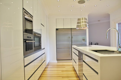 Photo of a large modern l-shaped open plan kitchen in Melbourne with an undermount sink, flat-panel cabinets, white cabinets, quartz benchtops, mirror splashback, stainless steel appliances, light hardwood floors and multiple islands.
