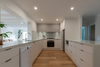 Photo of a large contemporary u-shaped open plan kitchen in Canberra - Queanbeyan with an undermount sink, flat-panel cabinets, white cabinets, quartz benchtops, glass sheet splashback, stainless steel appliances, light hardwood floors, no island, brown floor and white benchtop.