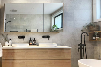 This is an example of a mid-sized contemporary master bathroom in Sydney with flat-panel cabinets, light wood cabinets, a freestanding tub, gray tile, ceramic tile, grey walls, ceramic floors, a vessel sink, solid surface benchtops, grey floor and white benchtops.