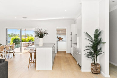 Design ideas for a mid-sized beach style galley eat-in kitchen in Wollongong with an undermount sink, flat-panel cabinets, white cabinets, quartz benchtops, white splashback, ceramic splashback, panelled appliances, light hardwood floors, with island and grey benchtop.