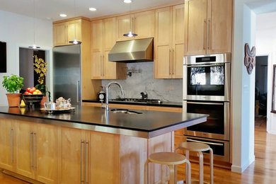 Example of a trendy u-shaped medium tone wood floor and brown floor eat-in kitchen design with a farmhouse sink, shaker cabinets, light wood cabinets, granite countertops, marble backsplash, stainless steel appliances, an island and black countertops