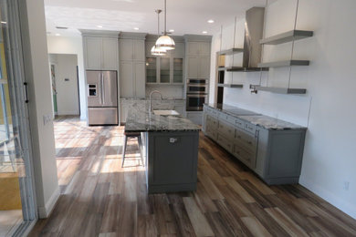 Mid-sized contemporary single-wall eat-in kitchen in Miami with a farmhouse sink, shaker cabinets, grey cabinets, granite benchtops, stainless steel appliances, medium hardwood floors, with island, brown floor and multi-coloured benchtop.