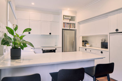 This is an example of a small contemporary open plan kitchen in Melbourne with a farmhouse sink, recessed-panel cabinets, white cabinets, quartz benchtops, white splashback, ceramic splashback, black appliances, medium hardwood floors, brown floor and white benchtop.