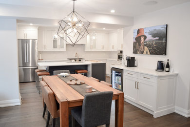 This is an example of a large contemporary kitchen/diner in Toronto with a double-bowl sink, shaker cabinets, white cabinets, engineered stone countertops, white splashback, engineered quartz splashback, stainless steel appliances, medium hardwood flooring, an island, brown floors and white worktops.