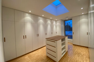 Geräumiges, Neutrales Modernes Ankleidezimmer mit Ankleidebereich, flächenbündigen Schrankfronten, weißen Schränken, hellem Holzboden und braunem Boden in Sonstige