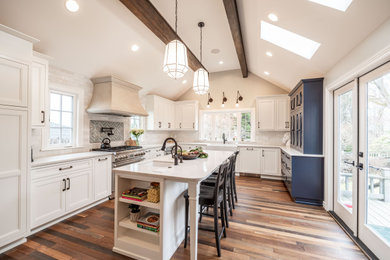 Transitional u-shaped medium tone wood floor, brown floor, exposed beam and vaulted ceiling kitchen photo in Seattle with a farmhouse sink, recessed-panel cabinets, white cabinets, quartz countertops, multicolored backsplash, subway tile backsplash, paneled appliances, an island and beige countertops