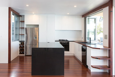 Inspiration for a mid-sized contemporary l-shaped open plan kitchen in Newcastle - Maitland with a drop-in sink, flat-panel cabinets, white cabinets, wood benchtops, white splashback, glass sheet splashback, black appliances, medium hardwood floors and with island.