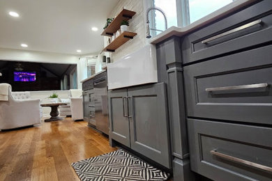 ルイビルにある高級な広いカントリー風のおしゃれなキッチン (エプロンフロントシンク、落し込みパネル扉のキャビネット、グレーのキャビネット、クオーツストーンカウンター、白いキッチンパネル、レンガのキッチンパネル、シルバーの調理設備、無垢フローリング、茶色い床、白いキッチンカウンター) の写真