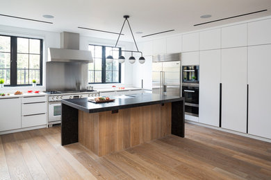 Inspiration for a contemporary l-shaped kitchen in London with flat-panel cabinets, white cabinets, metallic splashback, stainless steel appliances, light hardwood floors, with island and white benchtop.