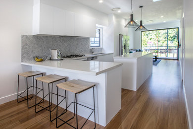 This is an example of a mid-sized modern l-shaped eat-in kitchen in Brisbane with a drop-in sink, flat-panel cabinets, white cabinets, granite benchtops, grey splashback, mosaic tile splashback, stainless steel appliances, light hardwood floors, multiple islands, white floor and white benchtop.