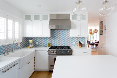 Example of a small beach style l-shaped light wood floor and brown floor enclosed kitchen design in Los Angeles with a farmhouse sink, shaker cabinets, blue backsplash, an island, white cabinets, solid surface countertops and ceramic backsplash