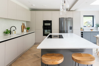 Medium sized modern grey and cream l-shaped kitchen/diner in Hampshire with flat-panel cabinets, white cabinets, white splashback, engineered quartz splashback, black appliances, light hardwood flooring, an island, white worktops, feature lighting, a submerged sink, engineered stone countertops and beige floors.