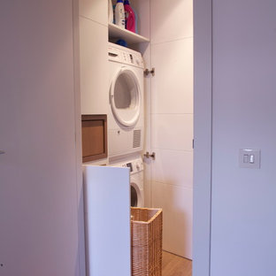 Diseño de armario lavadero actual, pequeño, con armarios con paneles lisos, puertas de armario blancas, paredes blancas, suelo de madera en tonos medios y lavadora y secadora apiladas