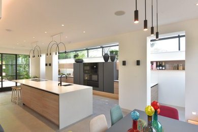 Inspiration for an expansive contemporary grey and brown u-shaped open plan kitchen in Manchester with a submerged sink, flat-panel cabinets, light wood cabinets, quartz worktops, white splashback, glass sheet splashback, black appliances, concrete flooring, an island, grey floors, white worktops and a vaulted ceiling.