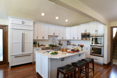 Large transitional l-shaped kitchen in Other with a farmhouse sink, white cabinets, quartz benchtops, white splashback, ceramic splashback, stainless steel appliances, medium hardwood floors, with island, brown floor, white benchtop, vaulted and shaker cabinets.