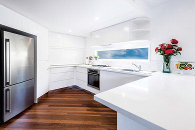 Design ideas for a mid-sized modern u-shaped kitchen in Sunshine Coast with flat-panel cabinets, white splashback, stone tile splashback, stainless steel appliances, medium hardwood floors, brown floor, white benchtop, a single-bowl sink, white cabinets and solid surface benchtops.