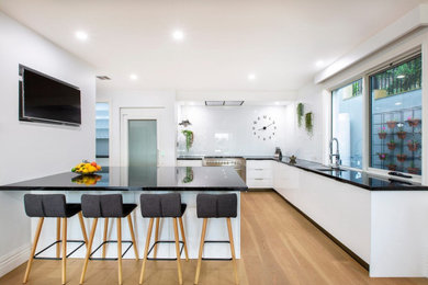 Design ideas for a large modern l-shaped open plan kitchen in Melbourne with a submerged sink, flat-panel cabinets, white cabinets, granite worktops, white splashback, glass sheet splashback, stainless steel appliances, light hardwood flooring, an island and black worktops.