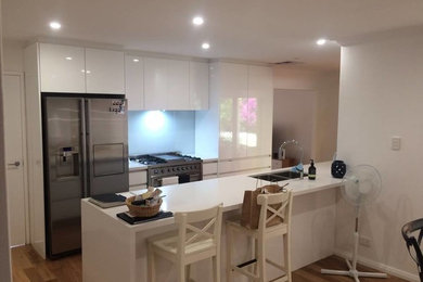Photo of a contemporary single-wall eat-in kitchen in Perth with a double-bowl sink, flat-panel cabinets, white cabinets, solid surface benchtops, grey splashback, glass sheet splashback, stainless steel appliances, light hardwood floors and a peninsula.