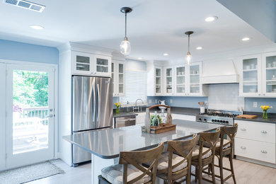 Mid-sized transitional l-shaped light wood floor and beige floor enclosed kitchen photo in Other with a farmhouse sink, flat-panel cabinets, white cabinets, solid surface countertops, white backsplash, subway tile backsplash, stainless steel appliances and an island
