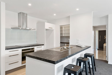 Photo of a mid-sized contemporary single-wall eat-in kitchen in Christchurch with a double-bowl sink, flat-panel cabinets, white cabinets, quartz benchtops, white splashback, stainless steel appliances, with island, black floor and black benchtop.