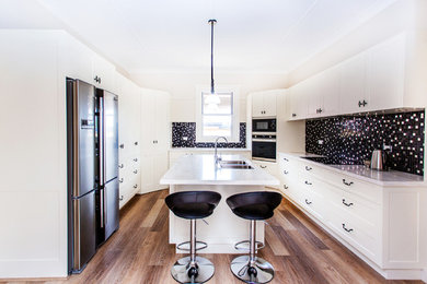 Mid-sized arts and crafts u-shaped eat-in kitchen in Other with an undermount sink, shaker cabinets, white cabinets, solid surface benchtops, black splashback, mosaic tile splashback, stainless steel appliances, medium hardwood floors, with island, brown floor and white benchtop.